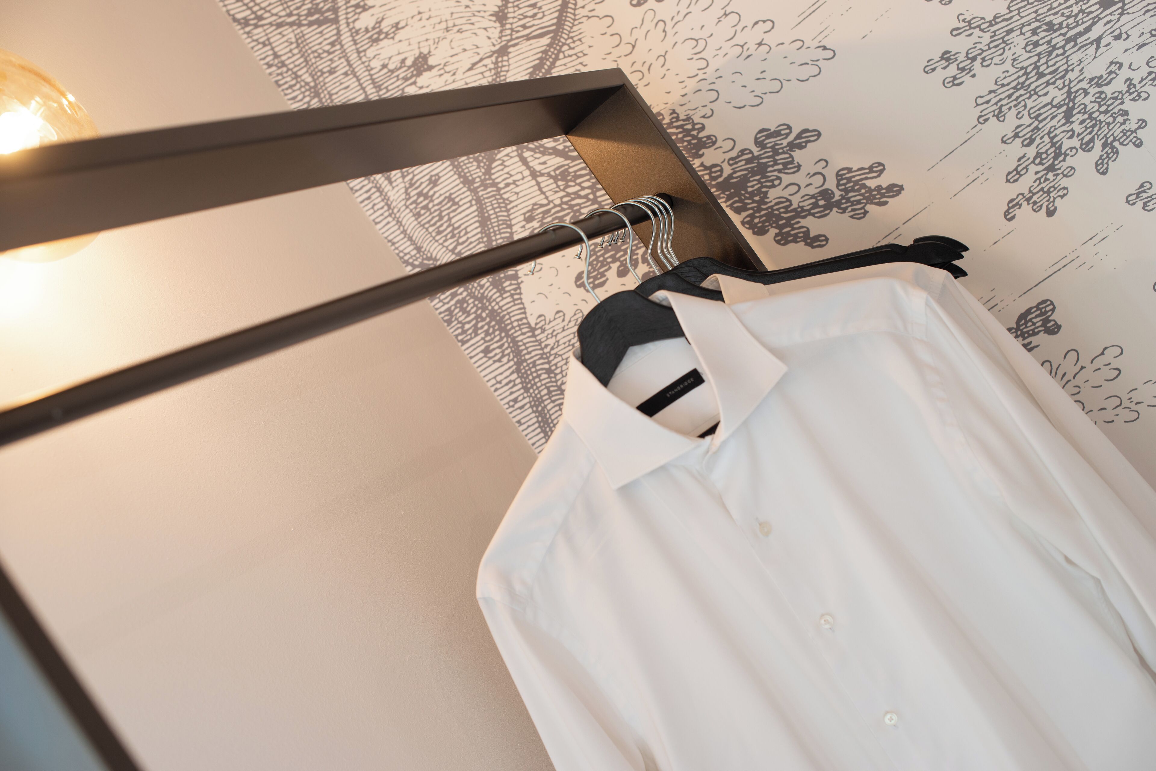 Una camisa blanca colgada en un perchero en una habitación con papel tapiz estampado y una lámpara visible.