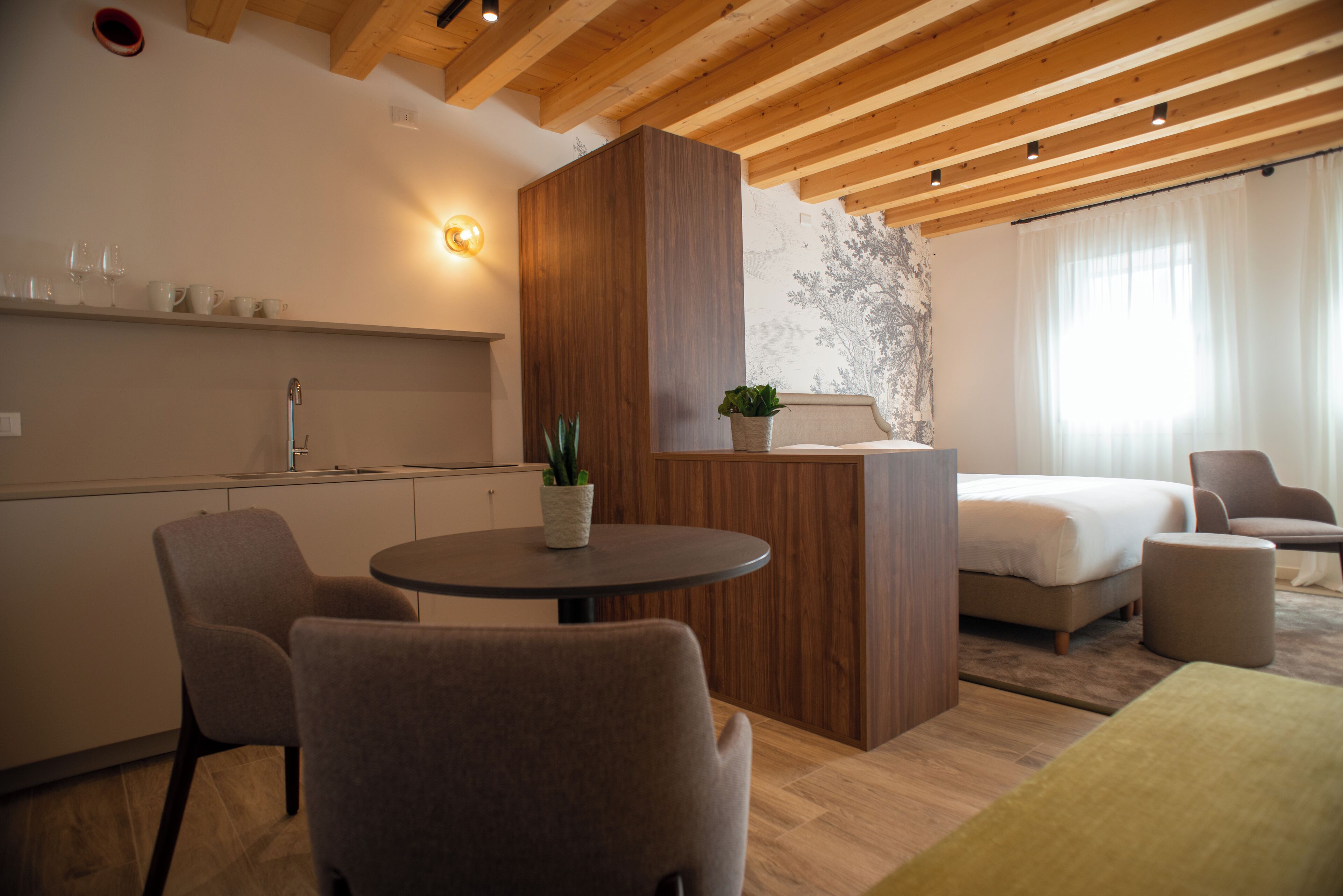 Una habitación de hotel moderna con techo de madera, una mesa redonda de comedor con sillas, una cama, una mesita auxiliar con una planta y un aplique de pared.