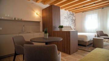 Una habitación de hotel moderna con techo de madera, una mesa redonda de comedor con sillas, una cama, una mesita auxiliar con una planta y un aplique de pared.