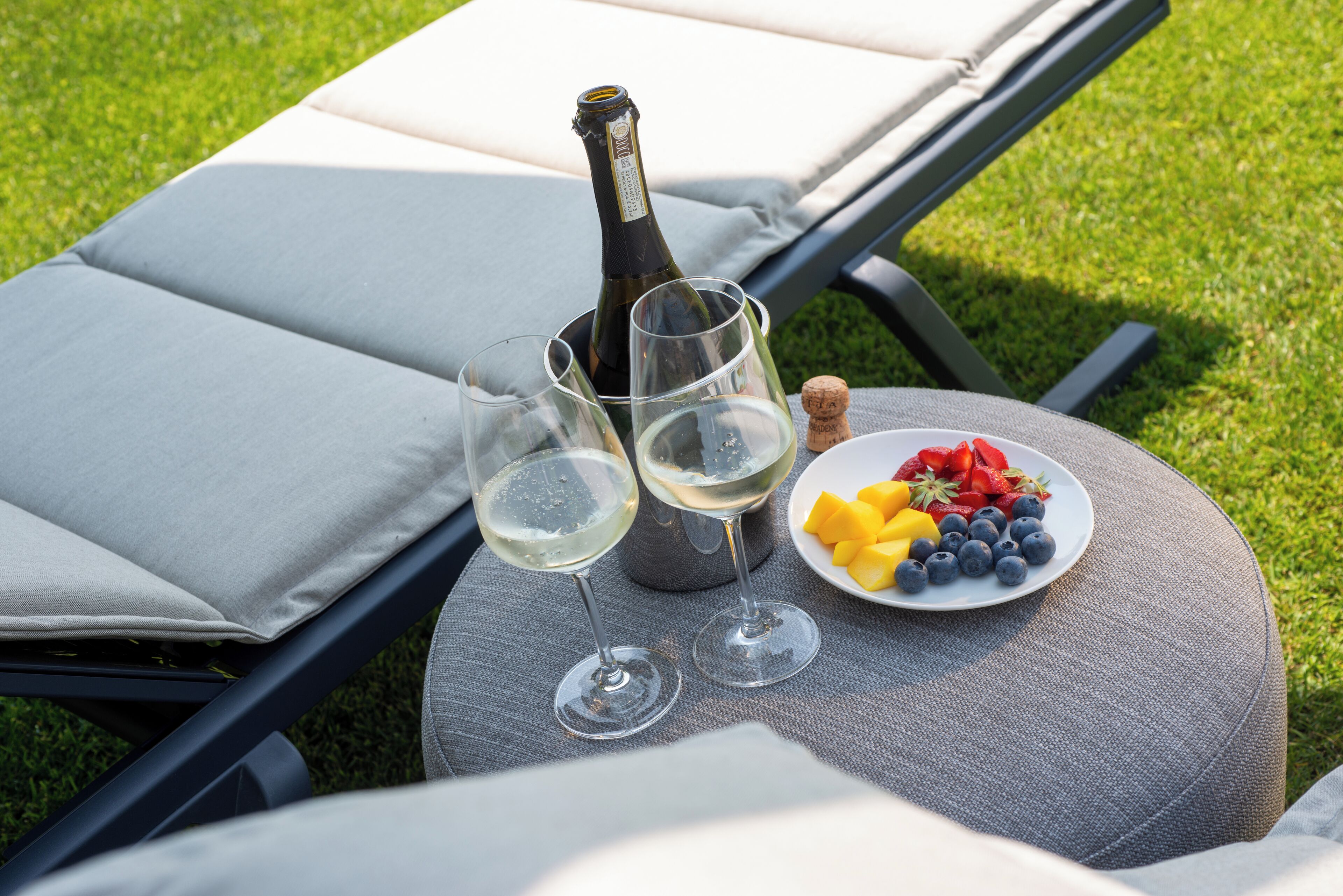 Eine Champagnerflasche, zwei Gläser und eine Obstplatte auf einem runden Tisch neben einem Liegestuhl.