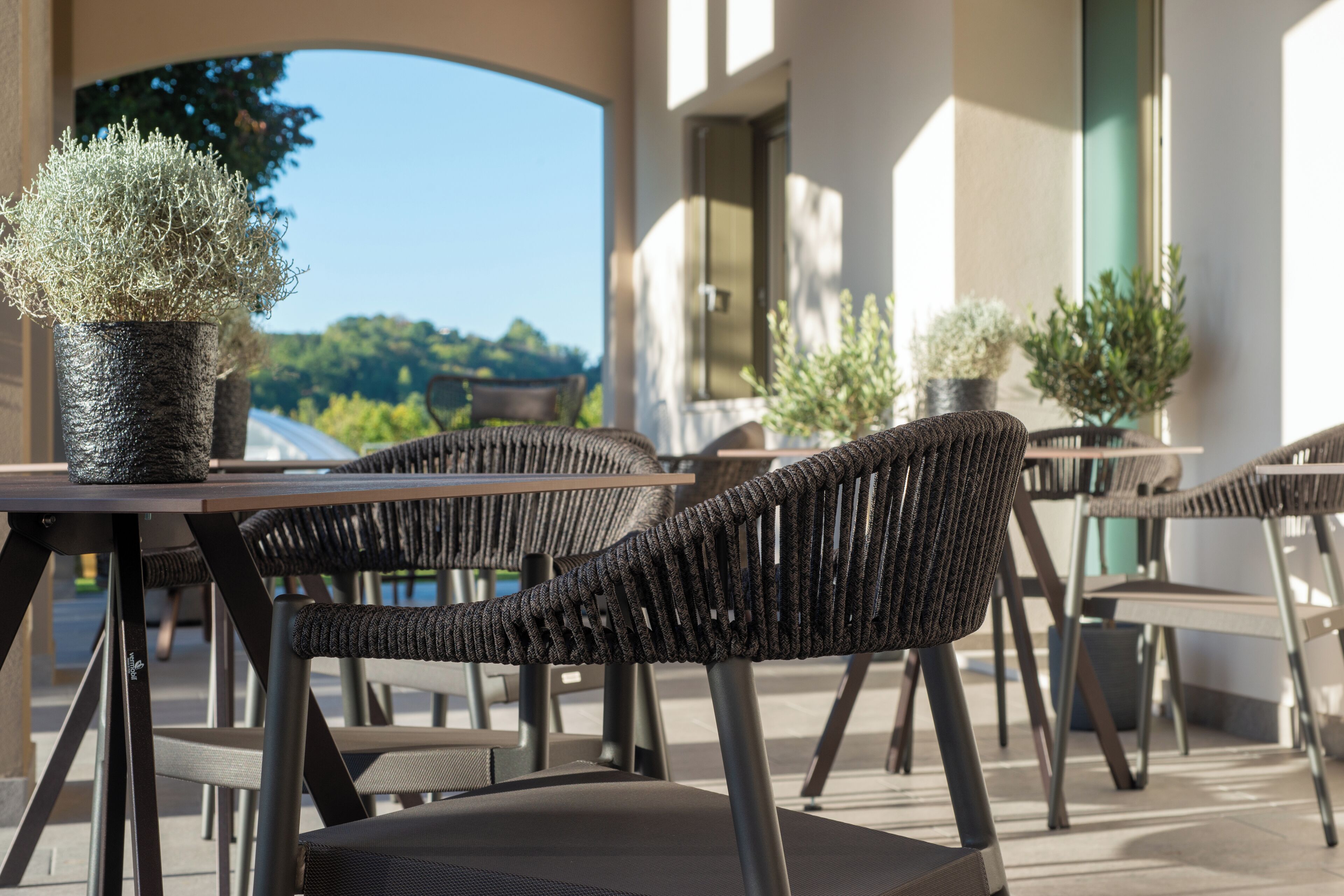 An outdoor seating area with wicker chairs and tables, potted plants, and a view of greenery in the background.