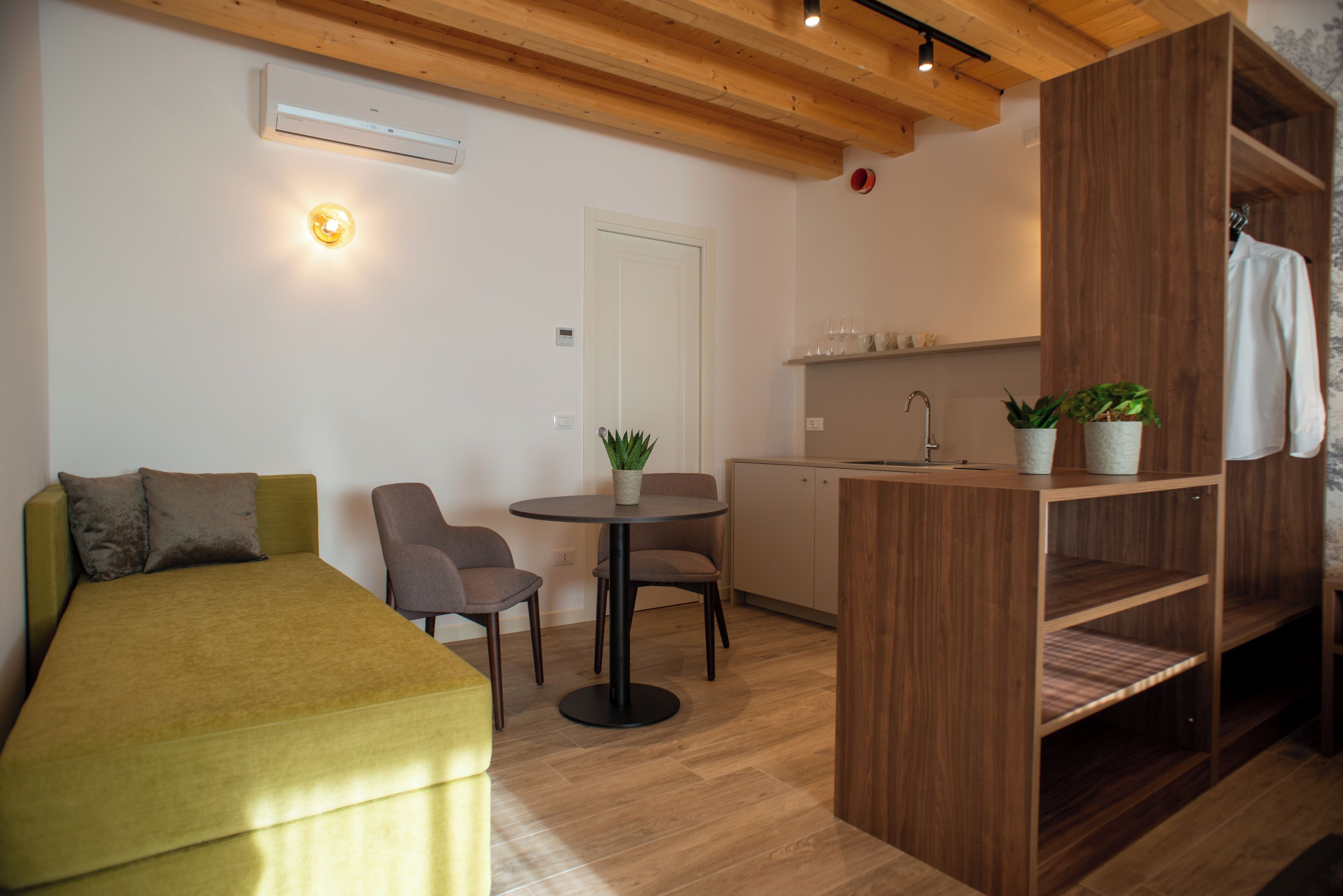 A compact room with a bed, a small table with chairs, a kitchenette, and a wooden shelf with plants.