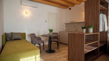A compact room with a bed, a small table with chairs, a kitchenette, and a wooden shelf with plants.