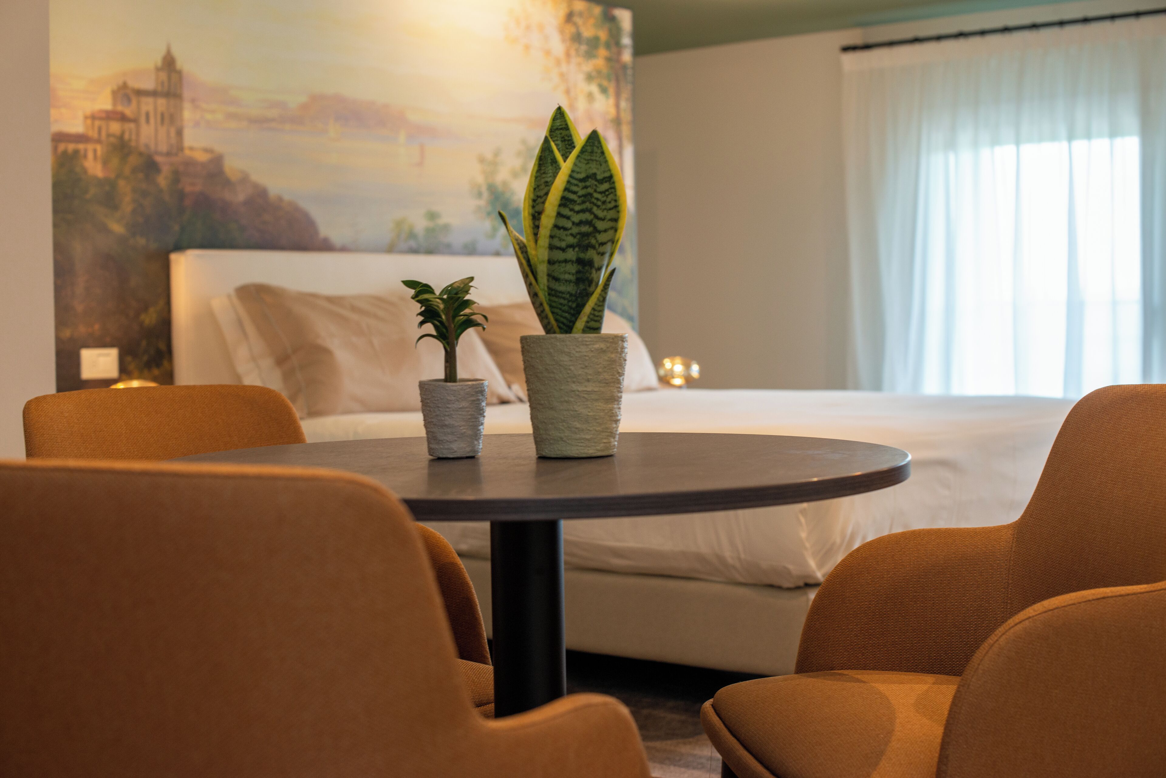 A hotel room with a bed, a round table with chairs, and two potted plants.