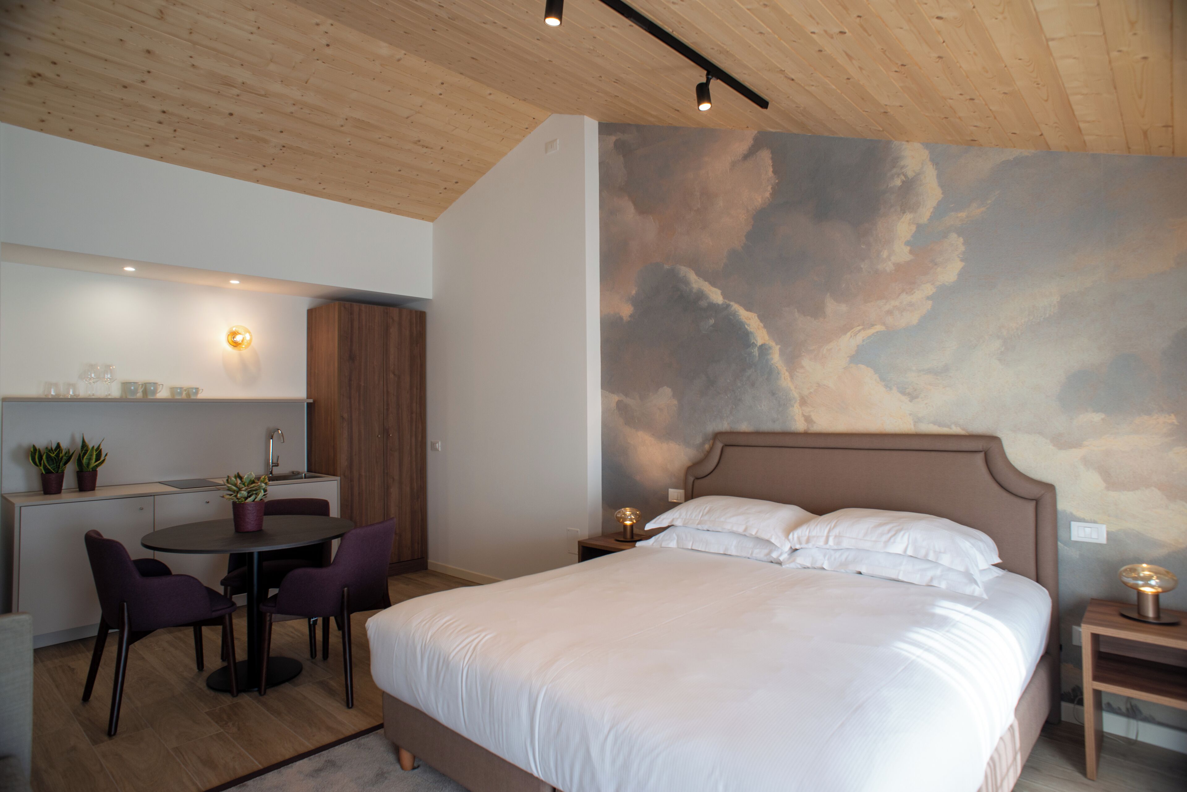 A bedroom with a large bed, a dining area with chairs and a table, and a wall with a cloud mural.