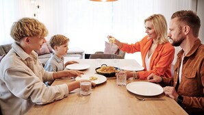 Uma família jantando à mesa de madeira, com pratos de comida e copos de água.