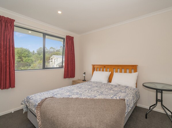 A bedroom with a wooden bed, a small bedside table, a nightstand, a window with red curtains, and a view of greenery outside. - Manuka Lodge - Tairua Holiday Home (Tairua)