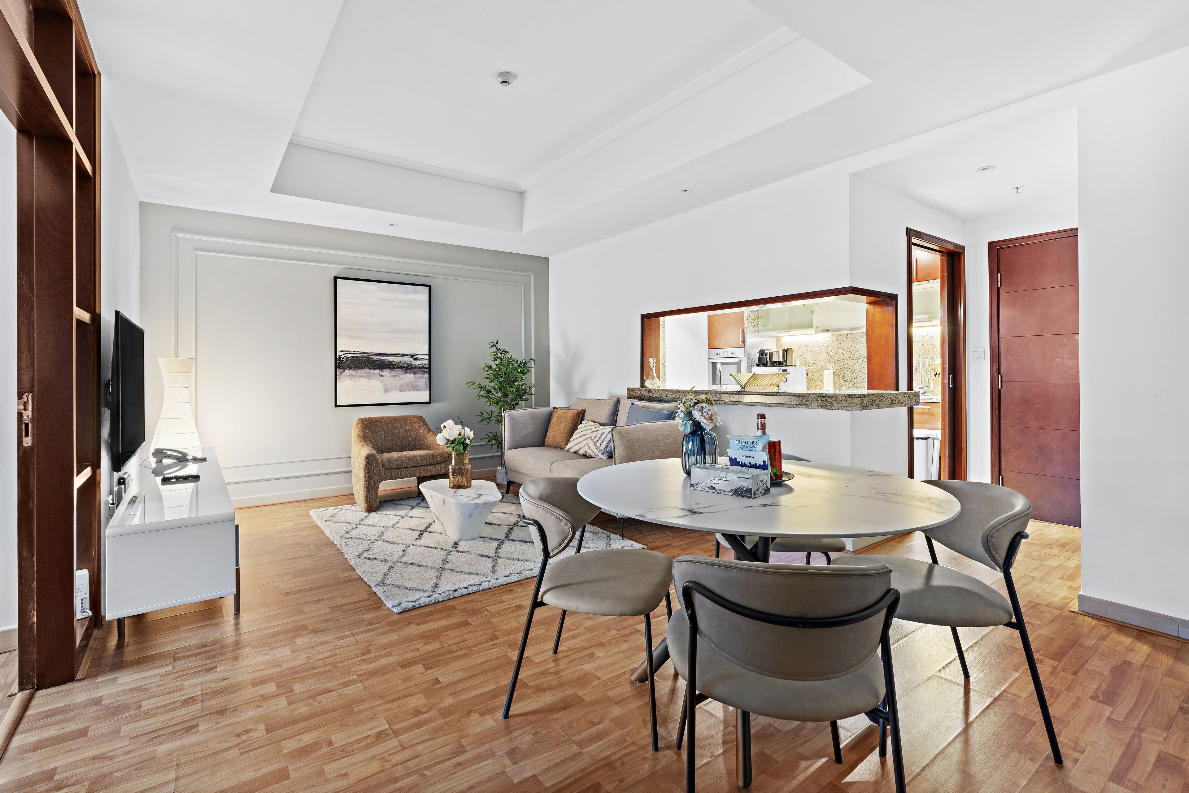 Un salon moderne avec une table à manger ronde, plusieurs chaises, un canapé, un téléviseur et un coin cuisine visible à travers une porte ouverte.