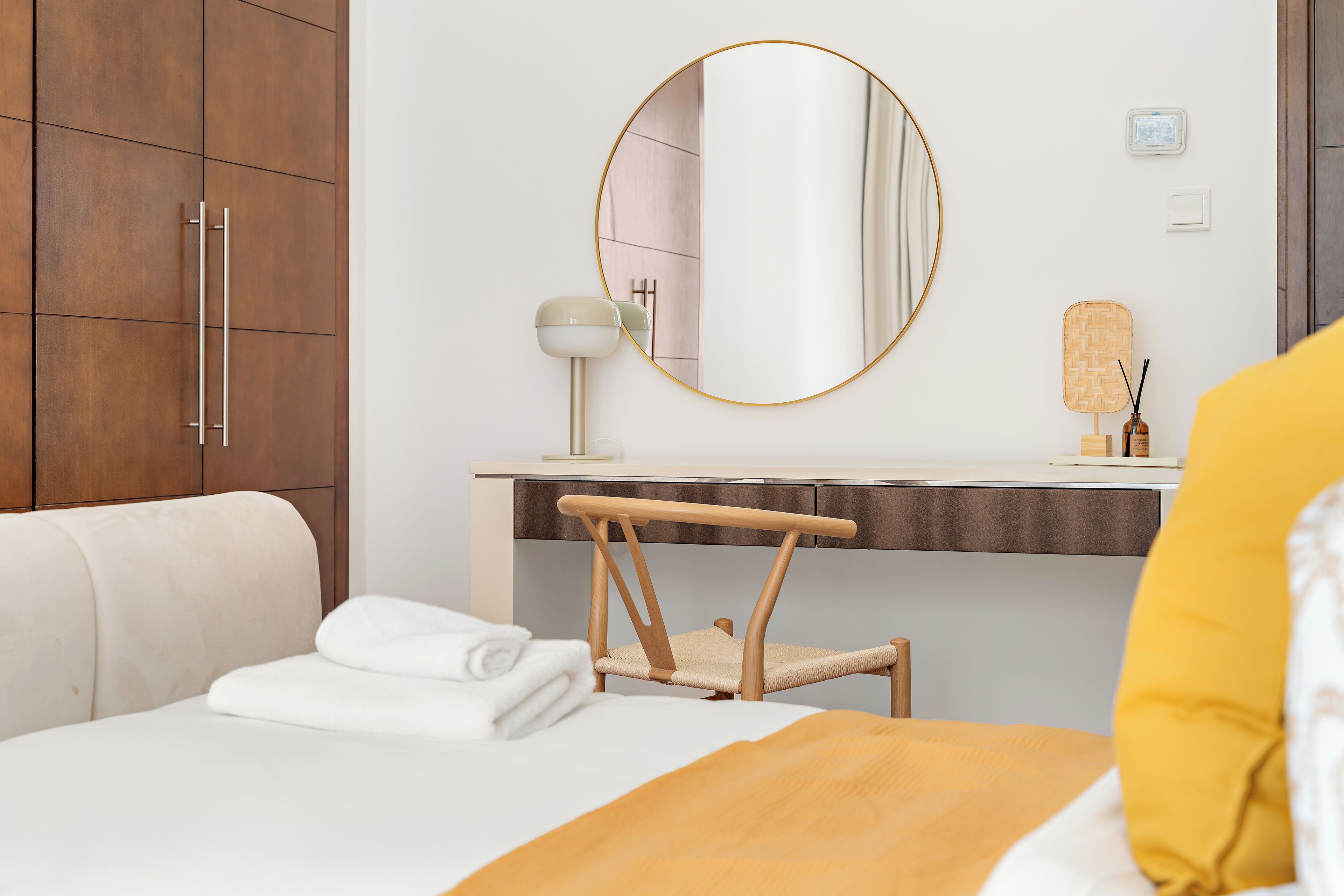 A hotel room with a bed, a wooden chair, a round mirror, a bedside table with a lamp, and a small shelf with decorative items.
