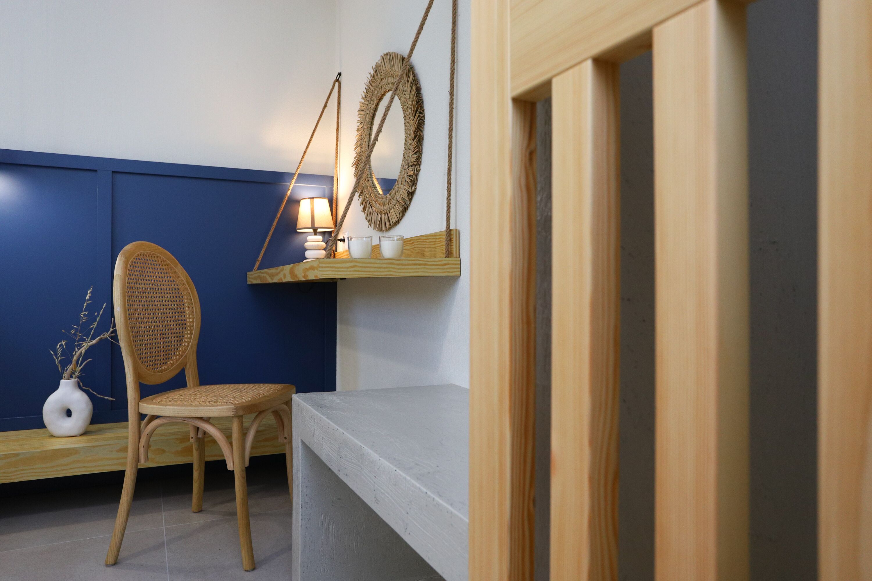 A room with a blue wall, a wooden chair, a white vase, a lamp, and a shelf with decorative items.