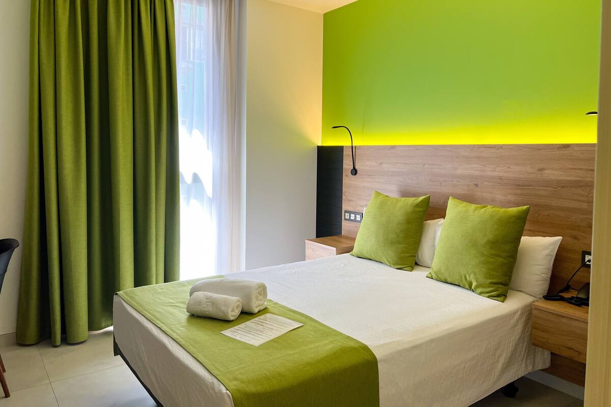 A hotel room with a bed, green curtains, a wooden headboard, and a bright wall.