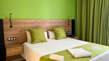 Habitación de hotel con cabecera de madera, paredes verdes, cama grande con ropa blanca y detalles verdes, mesita de noche con teléfono y toma de corriente, y cortina verde.