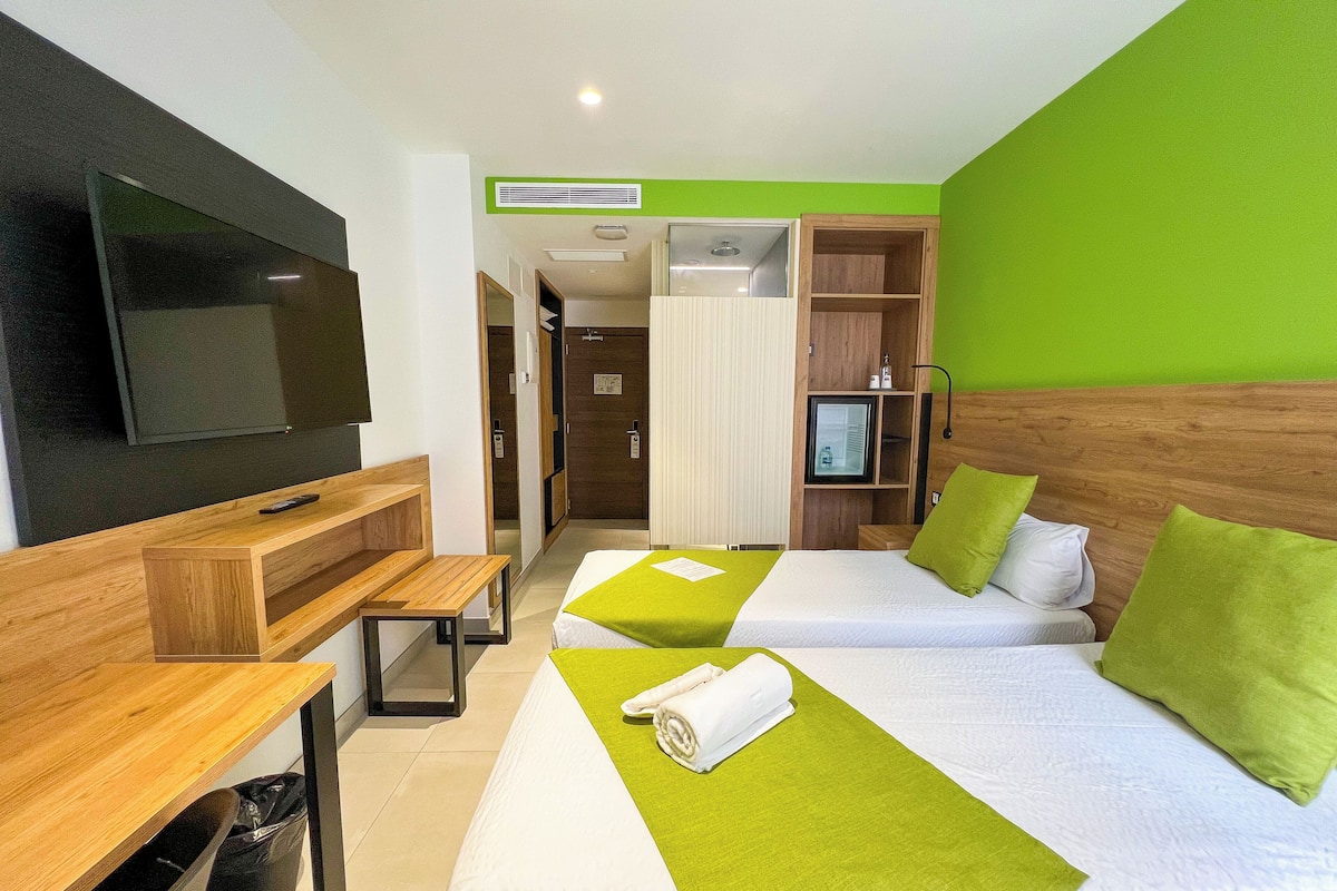A hotel room with two beds, a TV, a desk, and a green wall.