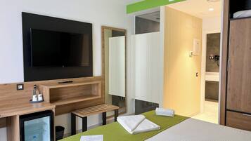 Habitación de hotel con un televisor de pantalla plana grande, un escritorio de madera con un refrigerador pequeño, una colcha verde y una cama bien hecha con toallas.