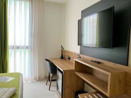 A modern hotel room with a wooden desk, a television mounted on the wall, a chair, and a bed with a green cover.