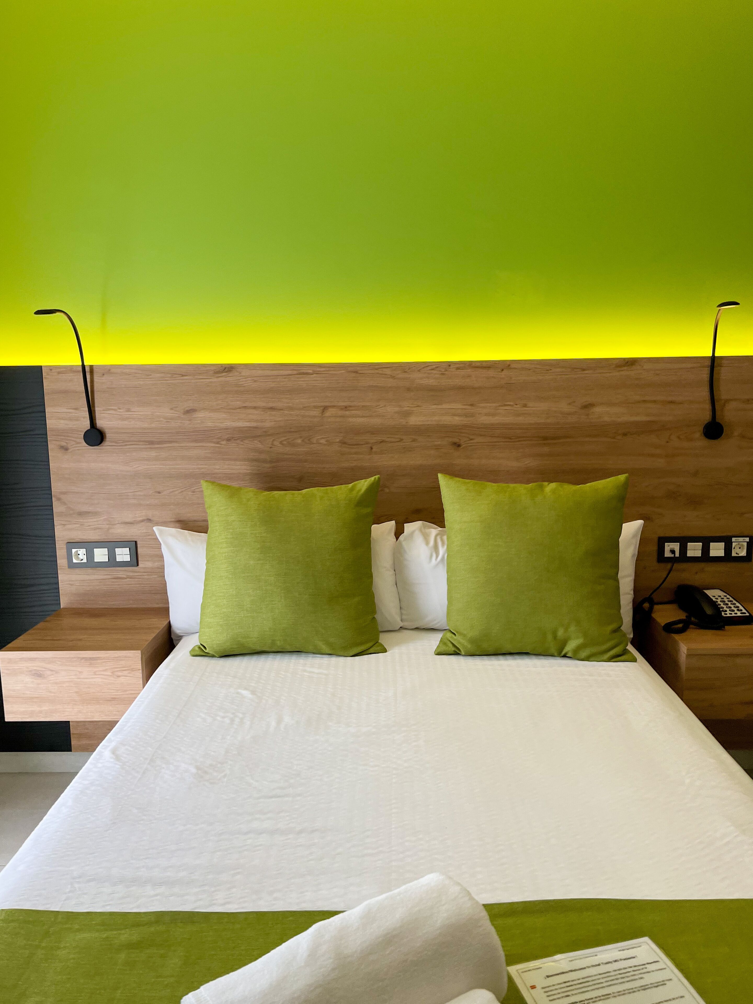 Ein Hotelzimmer mit einem hölzernen Kopfteil, einem Bett mit weißer Bettwäsche und zwei grünen Kissen, einem Nachttisch mit Telefon und einer leuchtend gelben Wand.