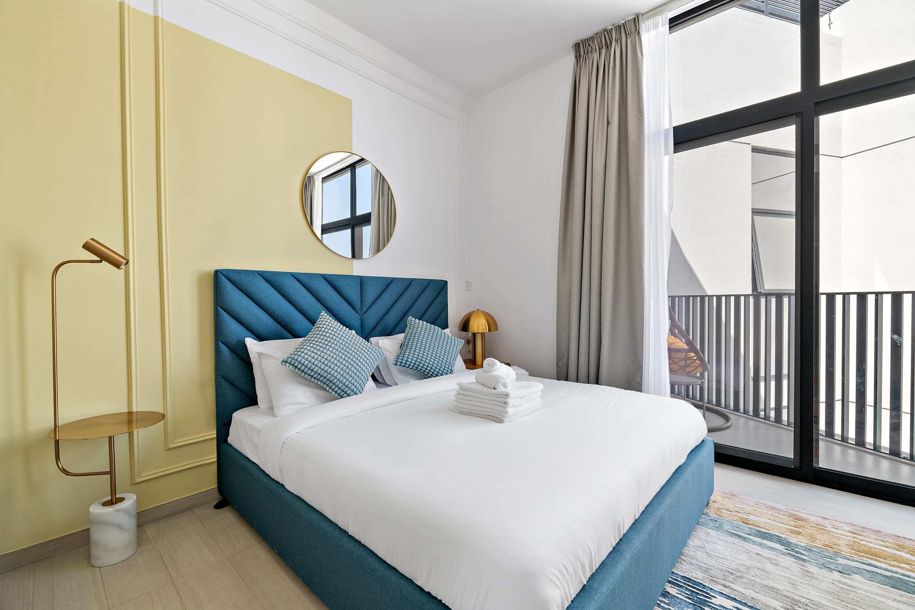A hotel room with a large bed, a round mirror, a bedside table with a lamp, and a view of the outside balcony.