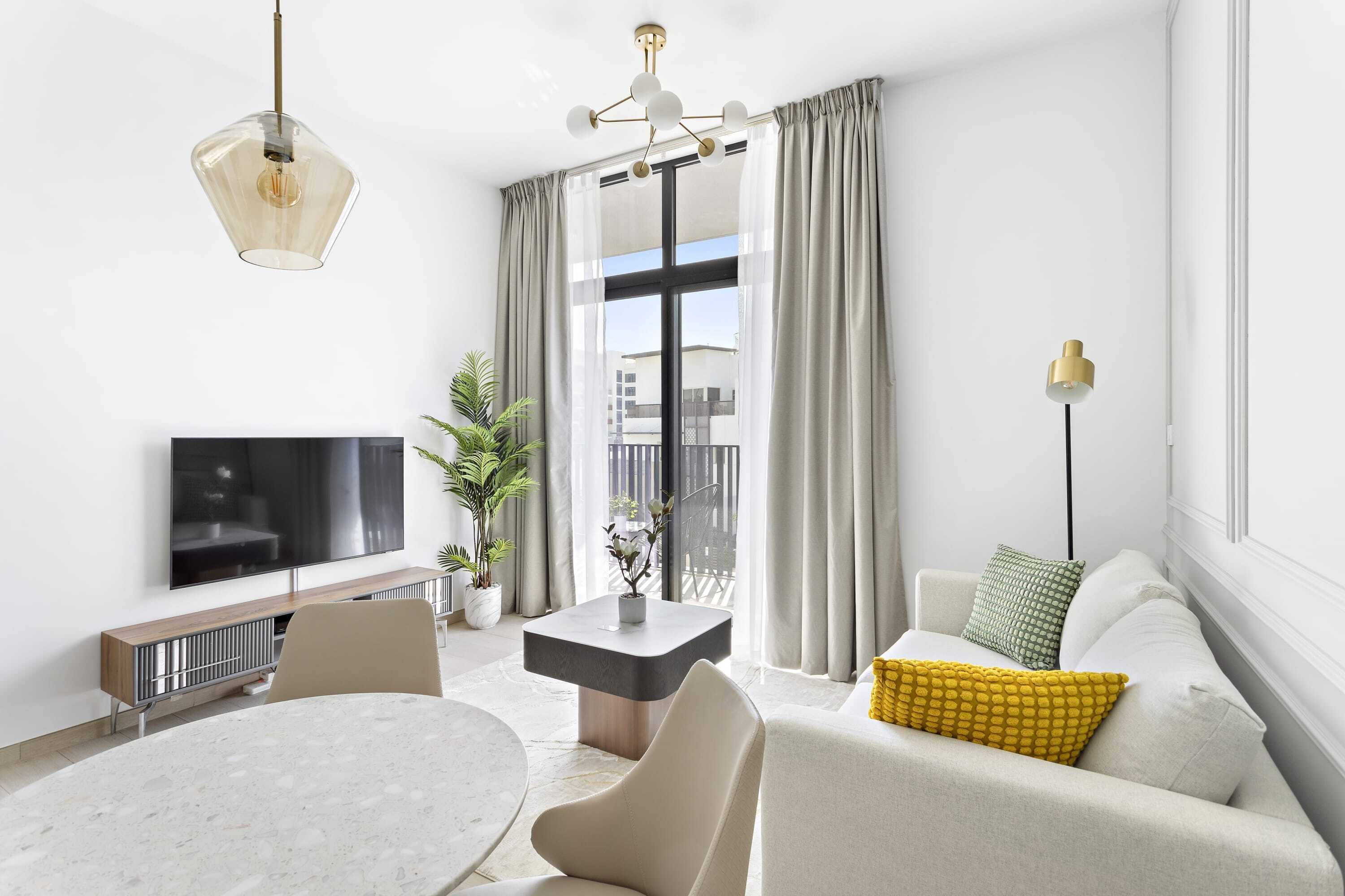 A modern living room with a flat-screen TV, a round dining table, a sofa with cushions, a side table, and a potted plant.