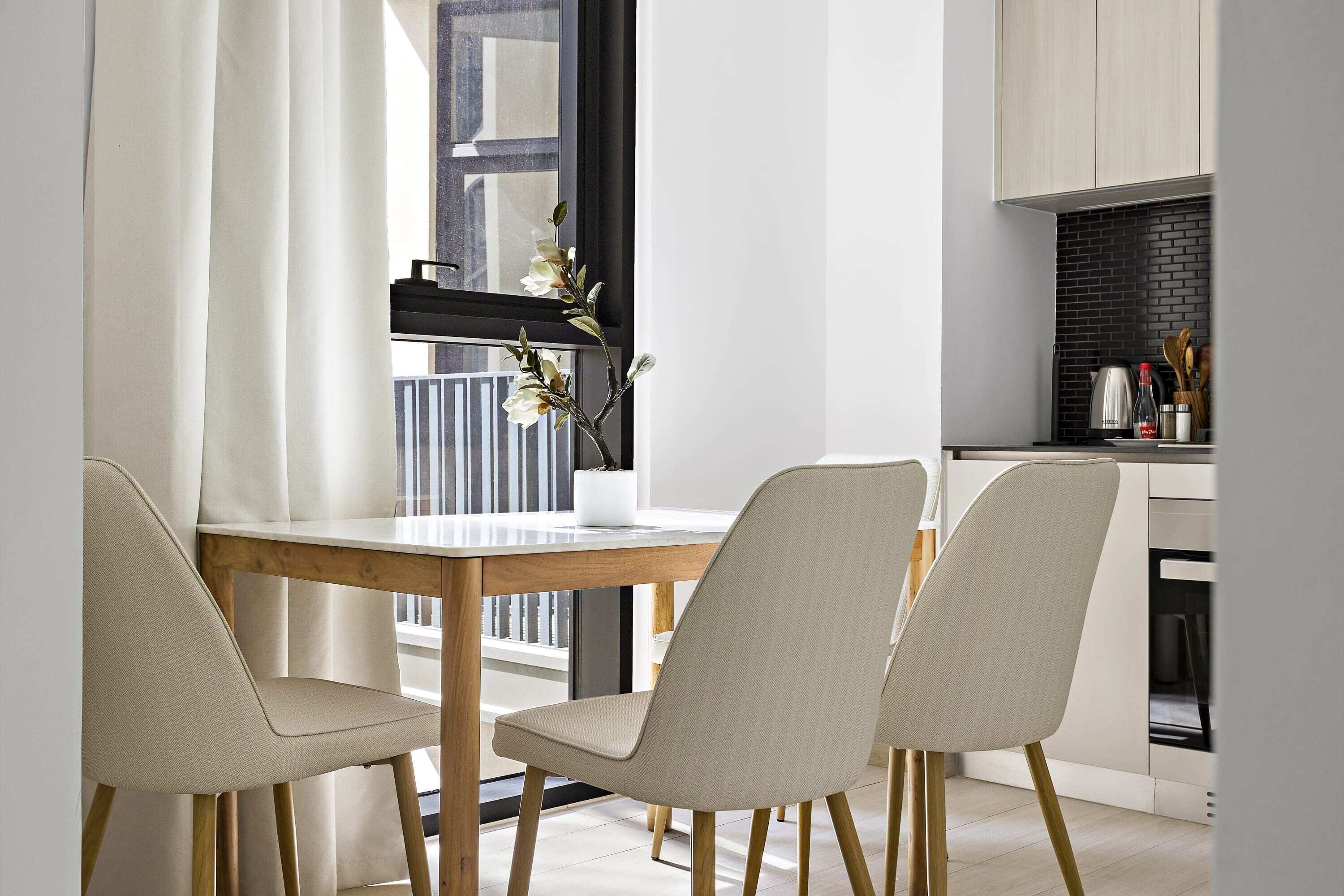 A modern dining area with a wooden table and two chairs, a vase with flowers, and a kitchen in the background.