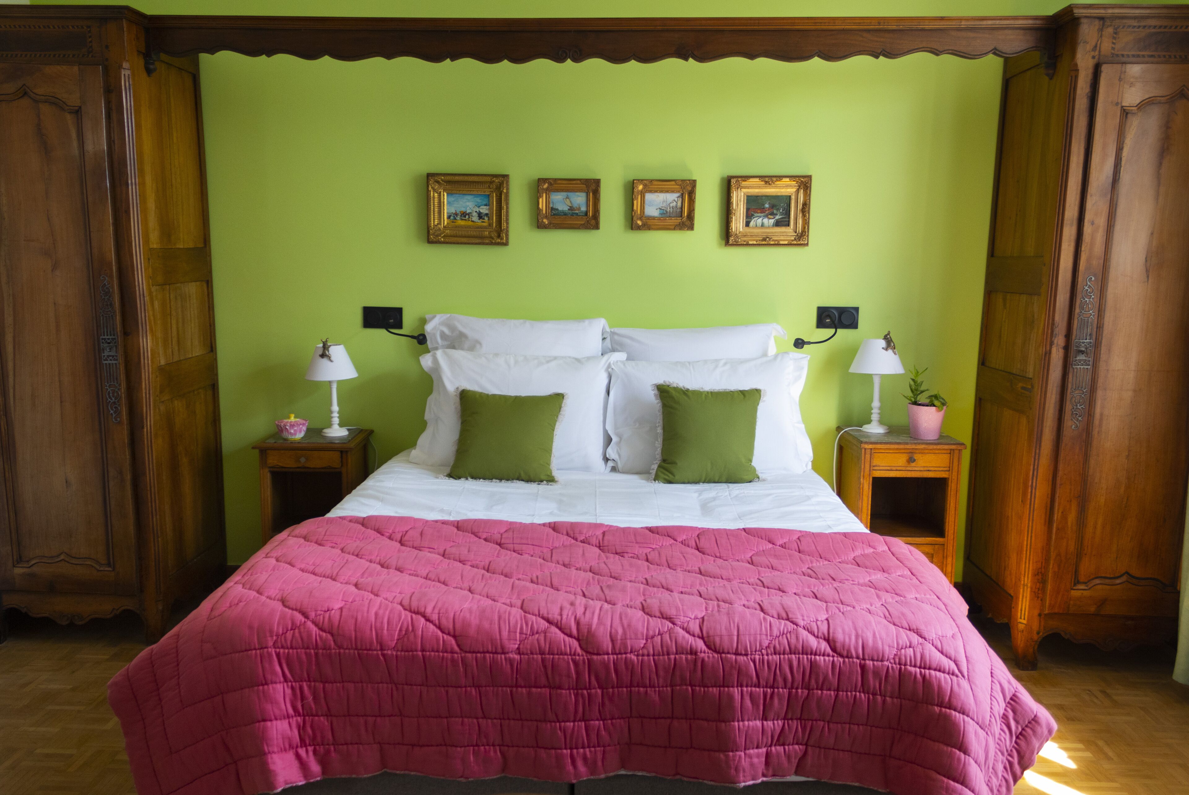 Un lit recouvert d’une couette rose, agrémenté d’oreillers blancs et de quelques oreillers décoratifs verts, avec de chaque côté des tables de chevet sur lesquelles reposent des lampes et un pot de fleurs.