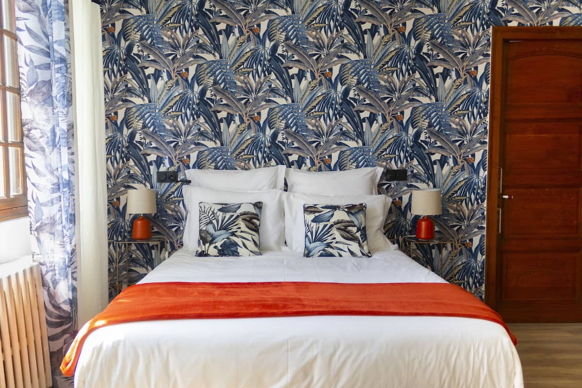A bed with white bedding, two pillows with a tropical leaf pattern, a red throw blanket, and a wall with a blue and white tropical leaf wallpaper.
