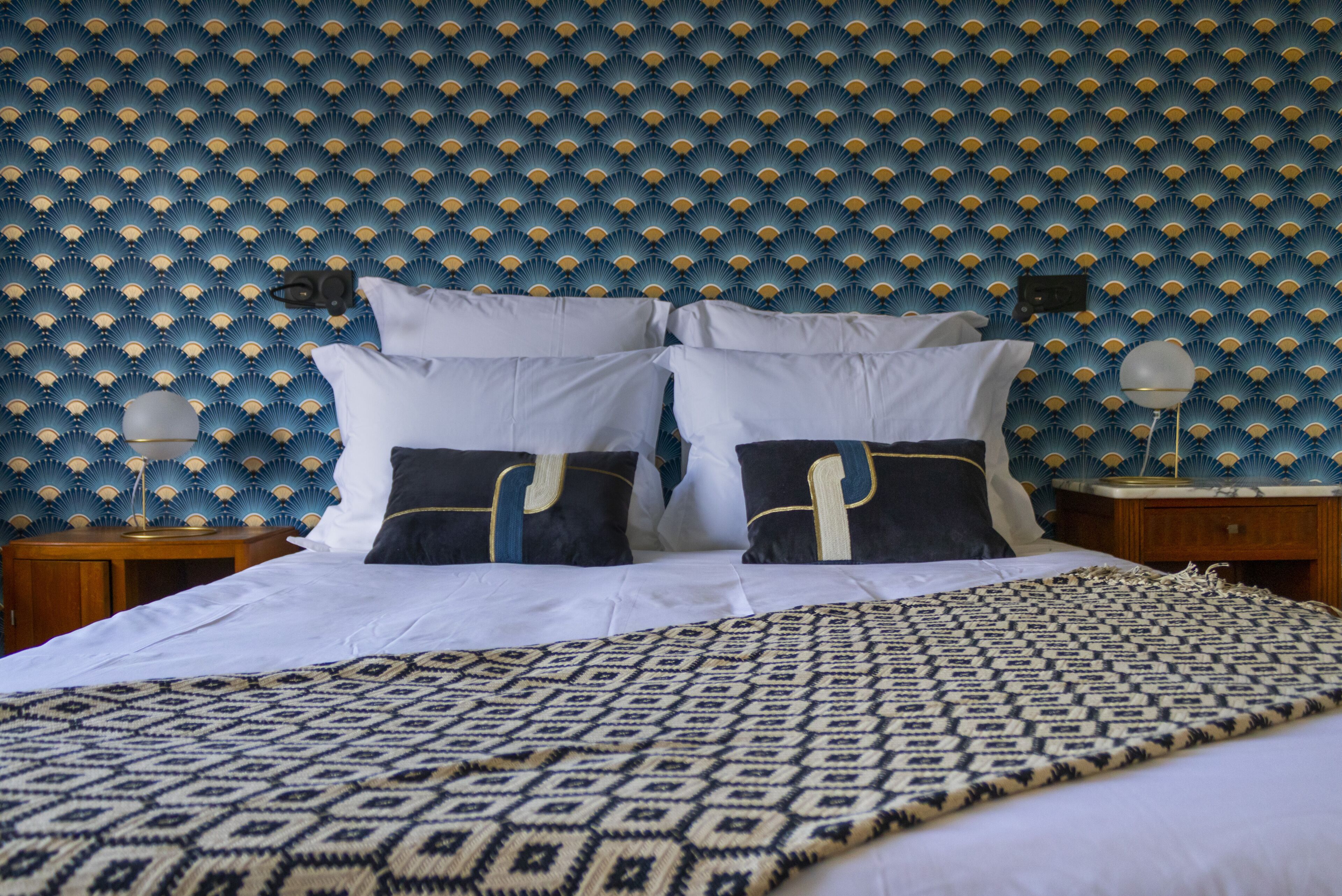 Una cama bien hecha con un edredón estampado, una pared con diseño azul y amarillo, y dos mesitas de noche con lámparas.