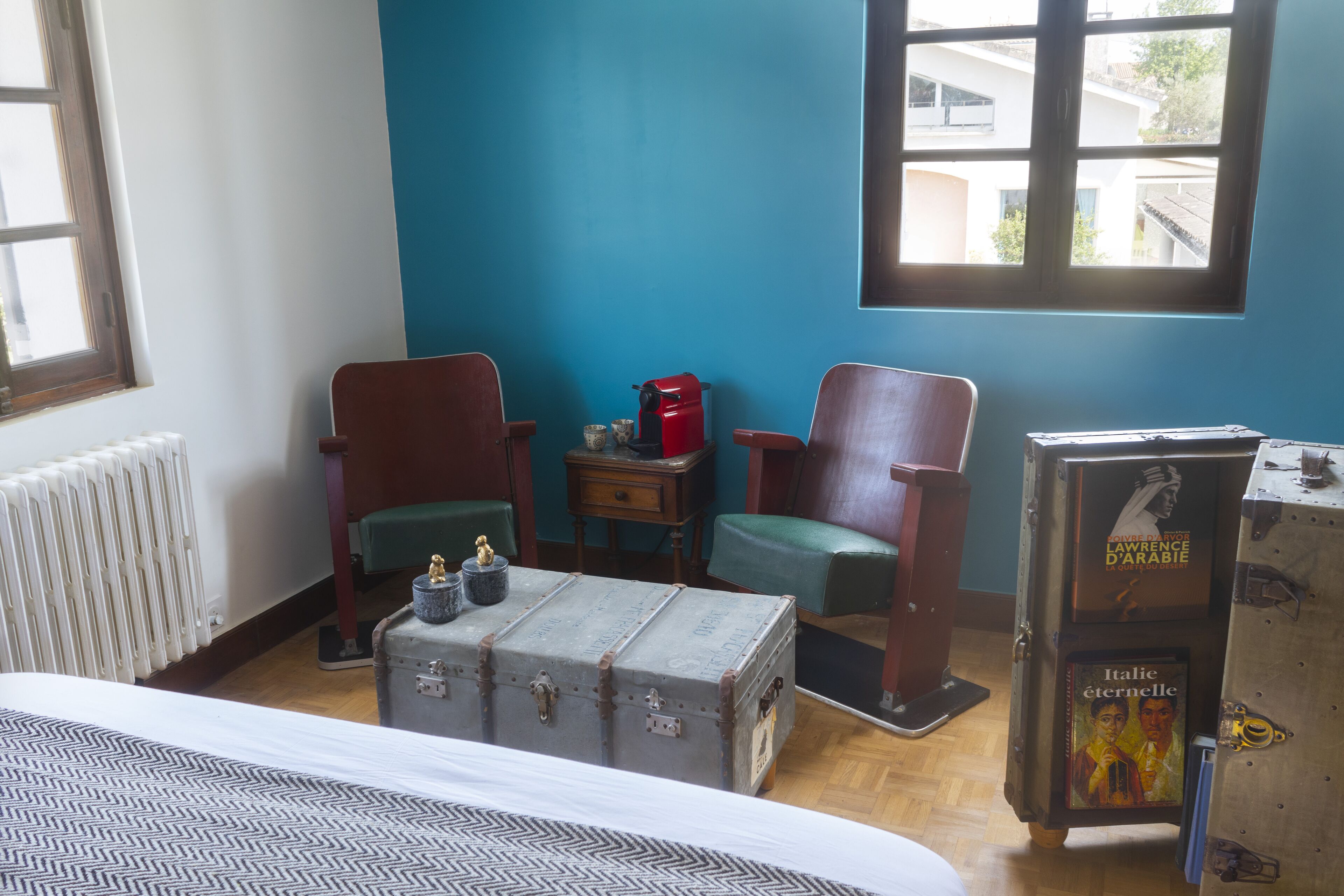 A room with a bed, two wooden chairs, a vintage suitcase, a radiator, and a window with a view of buildings.