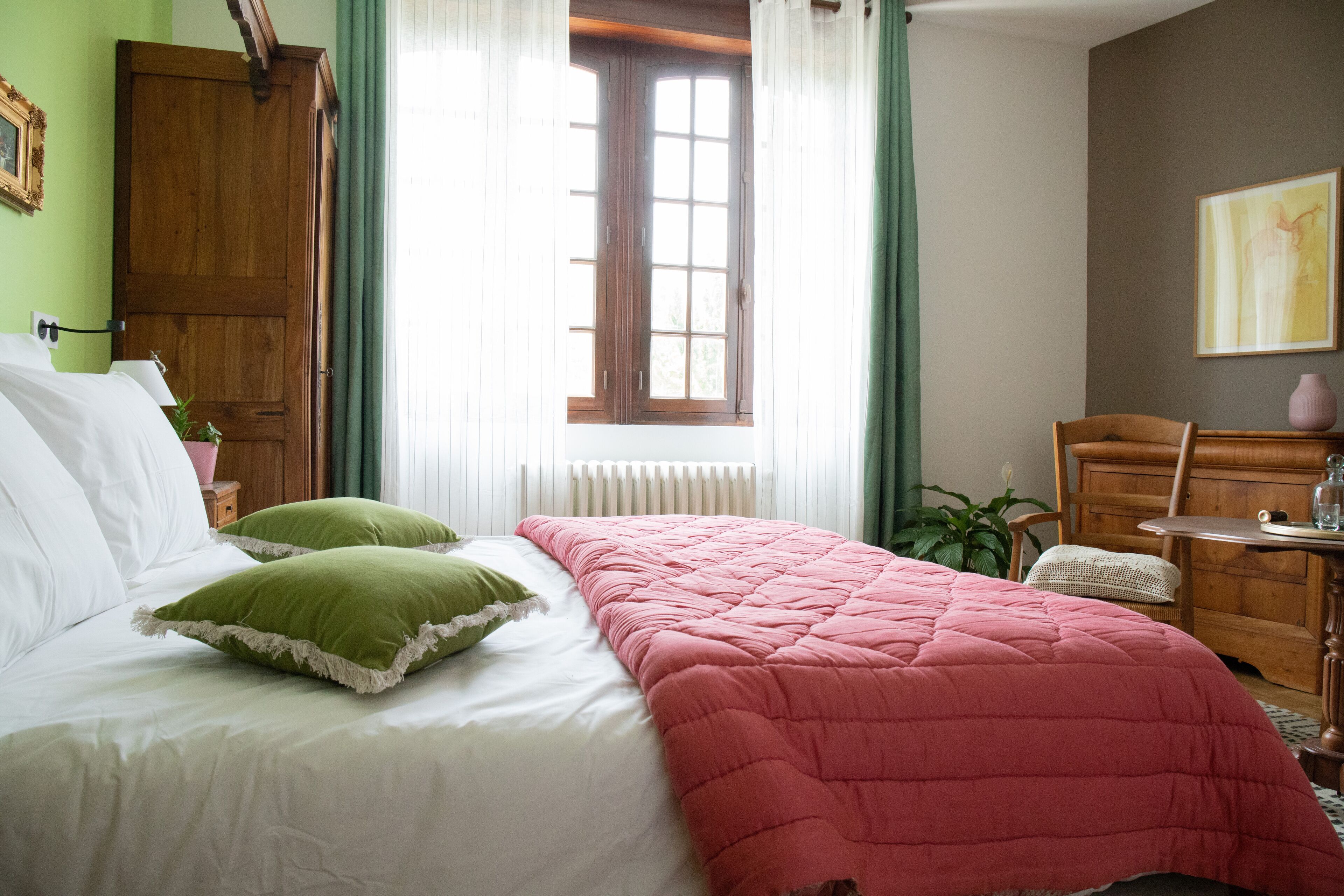 Une chambre à coucher avec un lit, des oreillers verts, une couverture rose, une armoire en bois, une fenêtre avec des rideaux, un bureau avec une chaise et un vase posé sur le bureau.