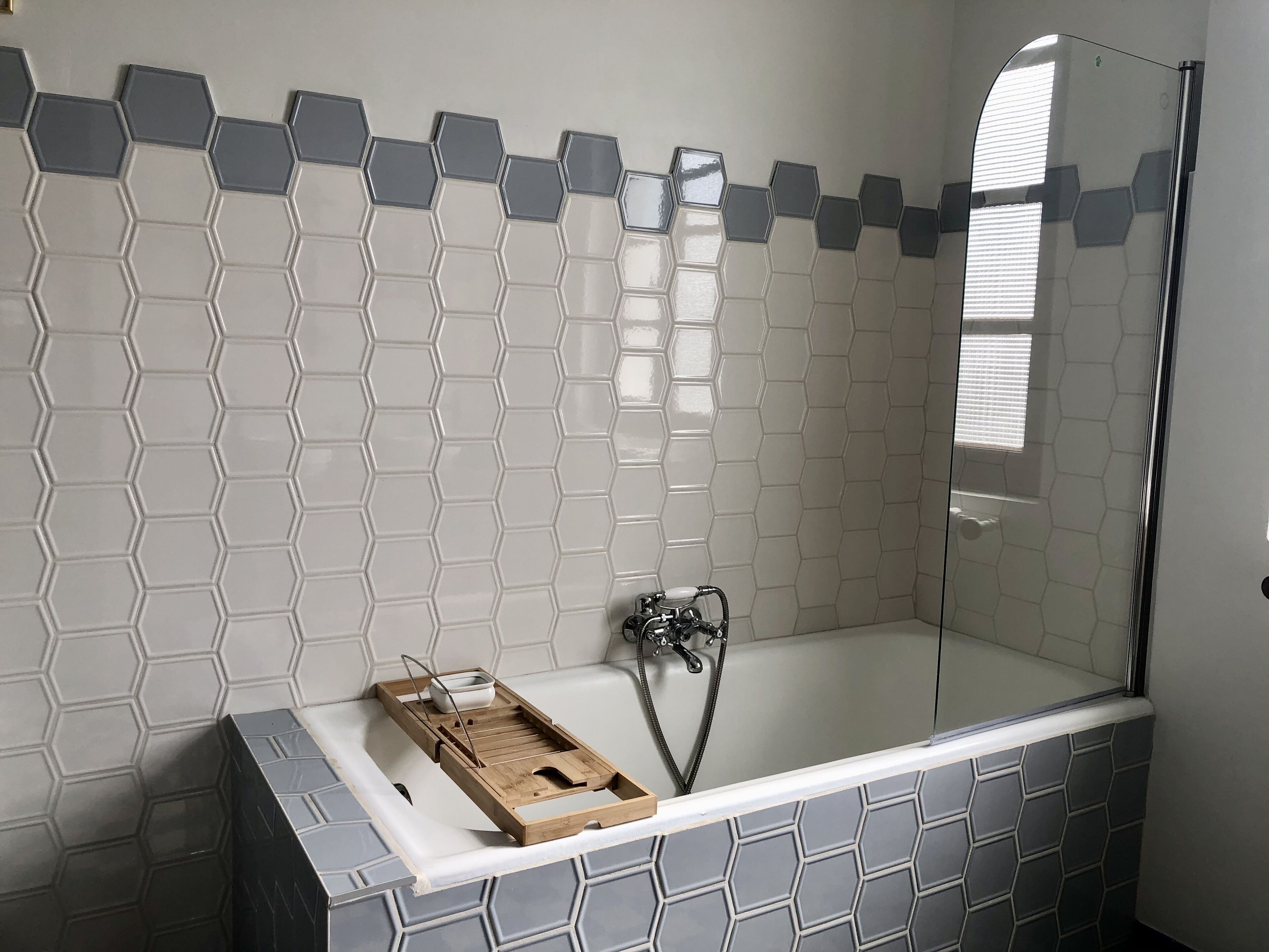 Une salle de bain moderne avec une douche à l’italienne en verre, un plateau en bois avec un bol, et une baignoire ornée de motifs hexagonaux.
