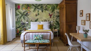 A bedroom with a bed, a wooden wardrobe, a desk with a chair, a potted plant, and a wall with a tropical mural. - Maison et table d'hôtes le Camélia Blanc (Saint-Ciers-sur-Gironde)