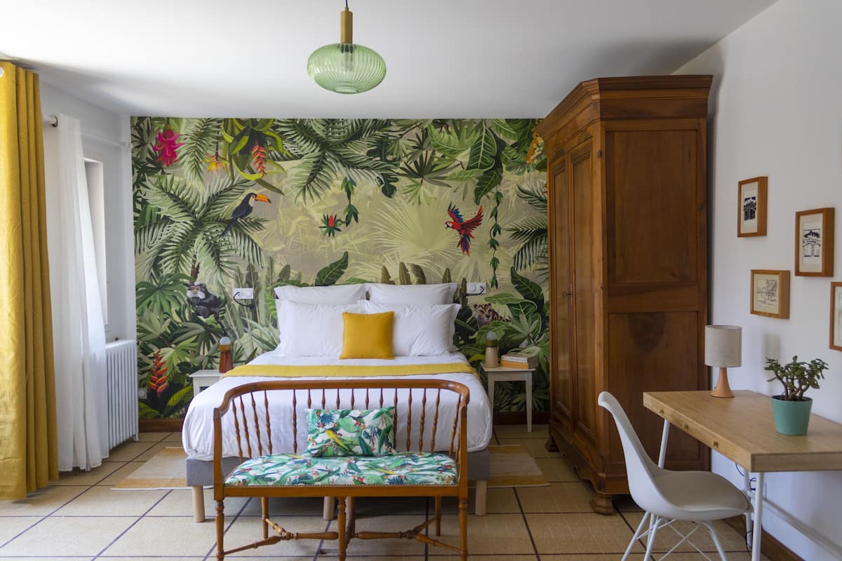 A bedroom with a bed, a wooden wardrobe, a desk with a chair, a potted plant, and a wall with a tropical mural.