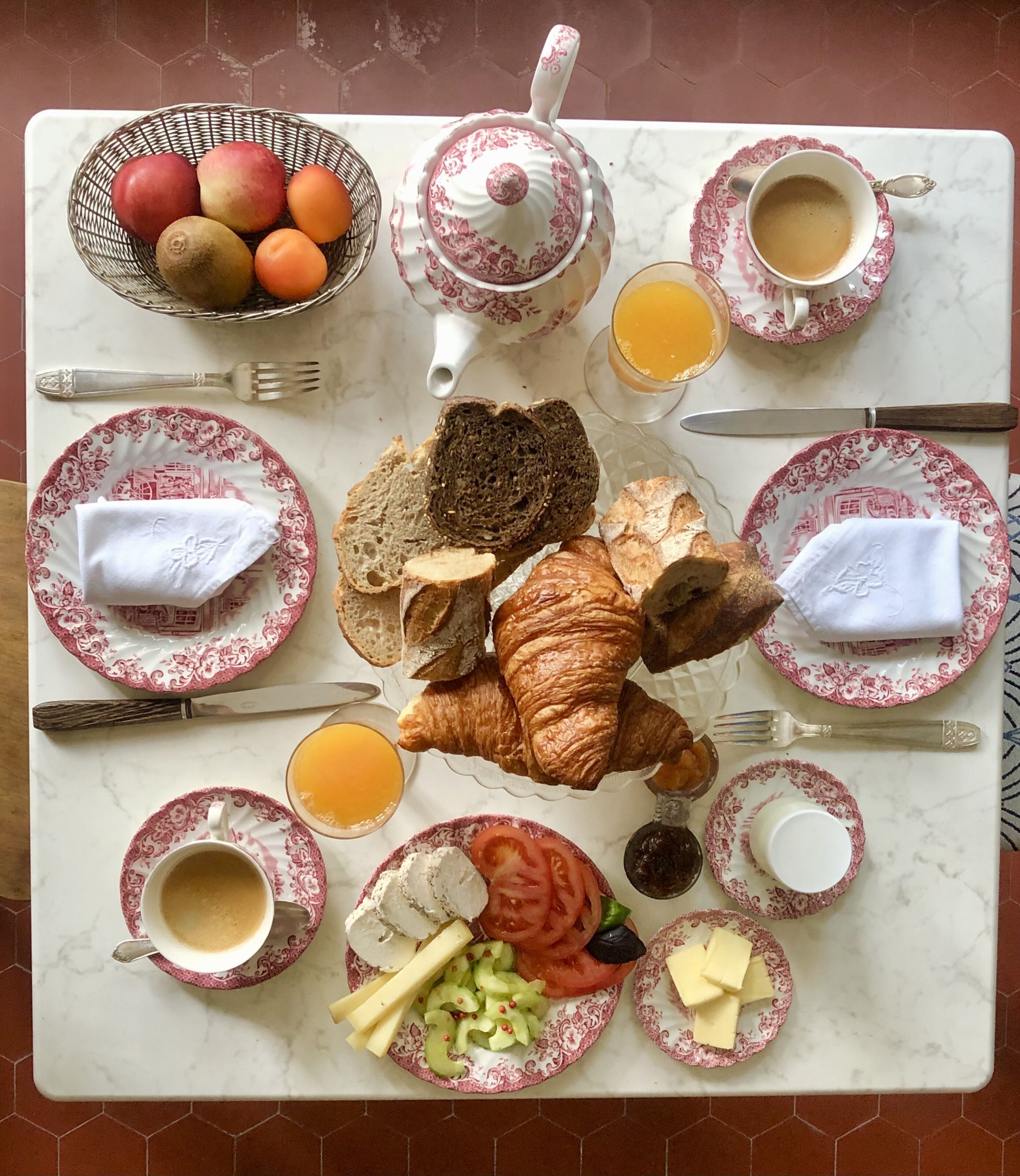 A breakfast table with croissants, bread, cheese, tomatoes, and a fruit bowl.