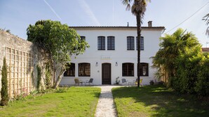 A white building with a central entrance, a paved pathway leading to the entrance, and a small garden area with chairs and plants. - Maison et table d'hôtes le Camélia Blanc (Saint-Ciers-sur-Gironde)