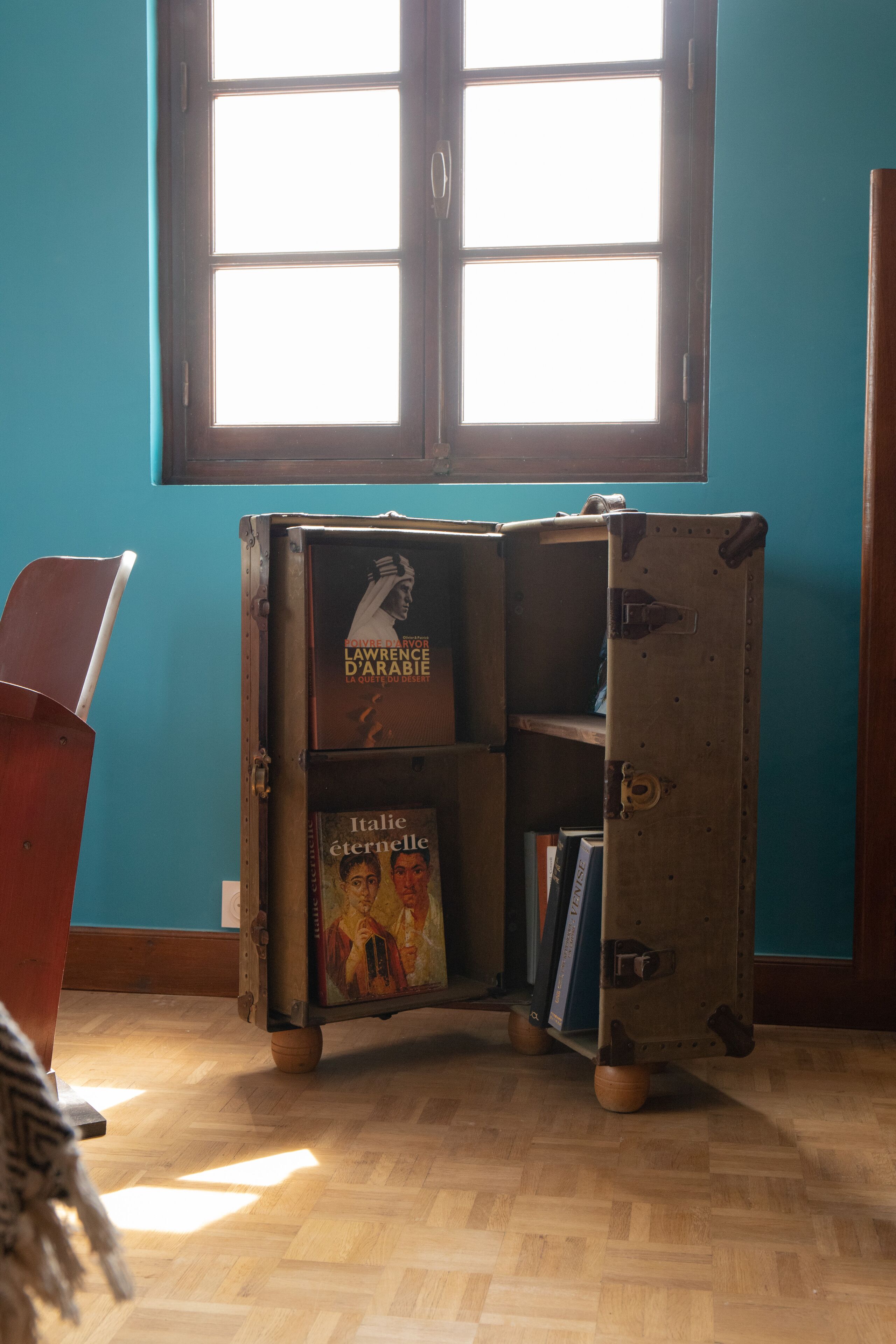 A room with a wooden floor, a window with blinds, and a vintage suitcase bookshelf containing books.