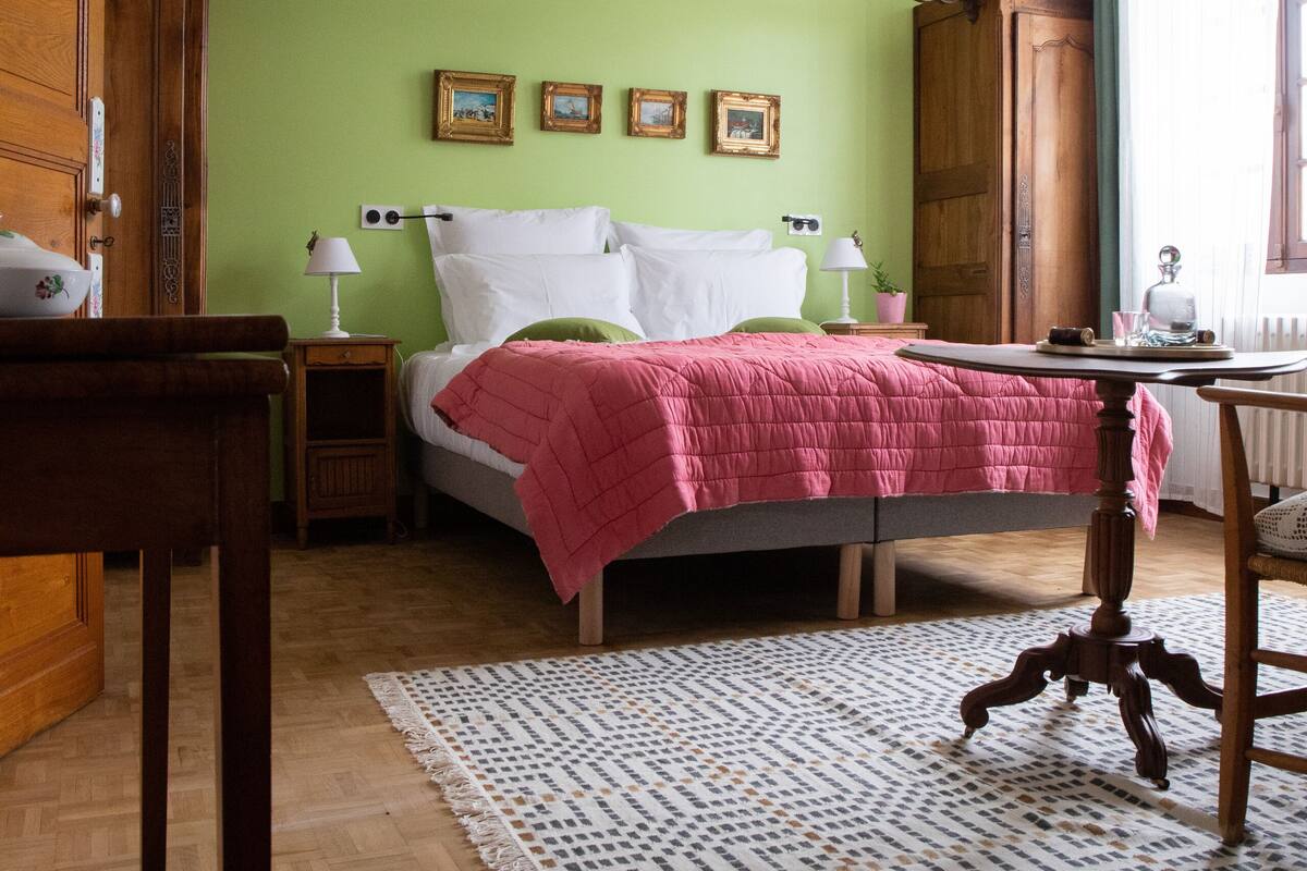A room with a bed, a wooden table, a chair, a lamp, and a patterned rug.