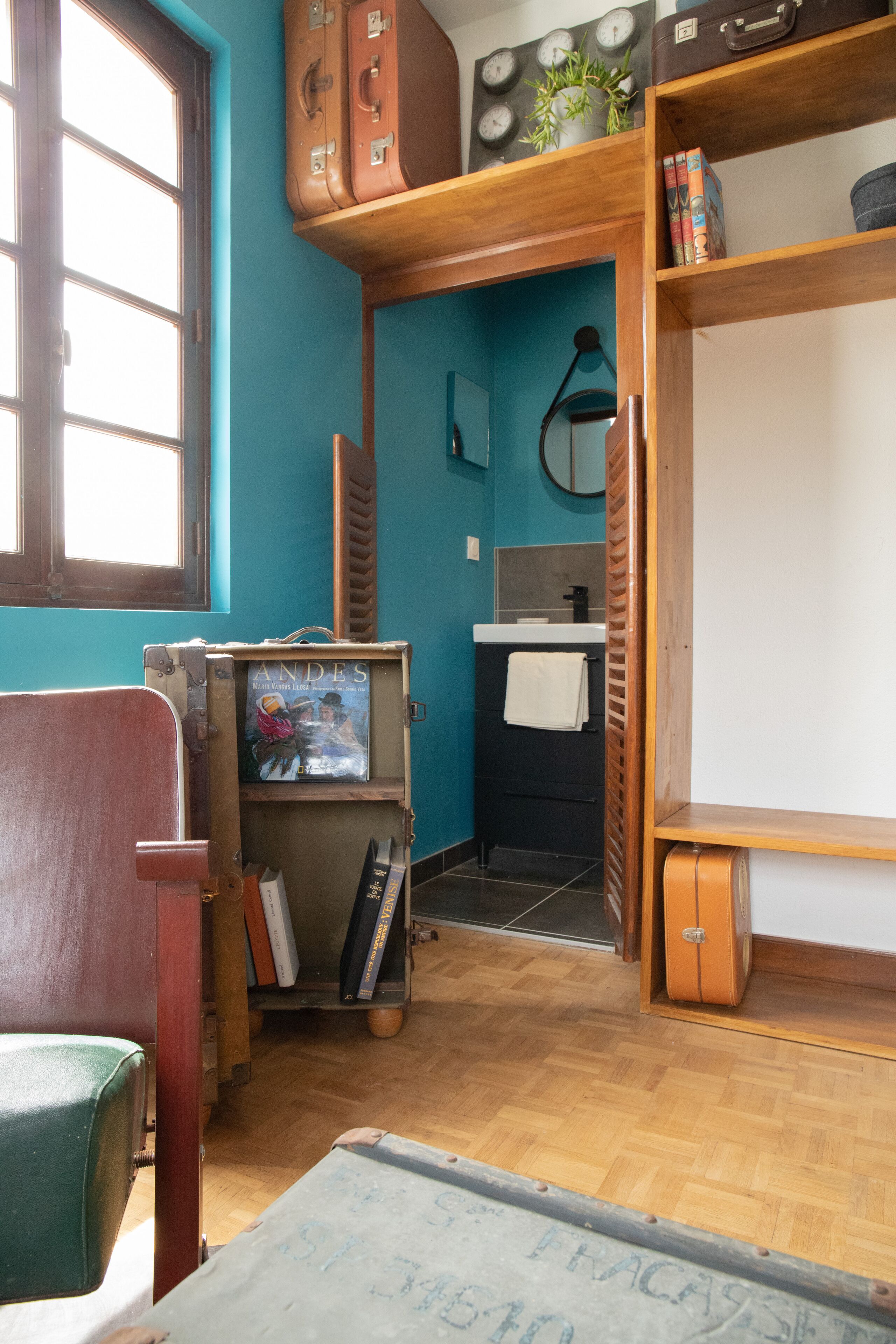 Una habitación con piso de madera, una ventana, un escritorio con una silla y una estantería con libros y una maleta.