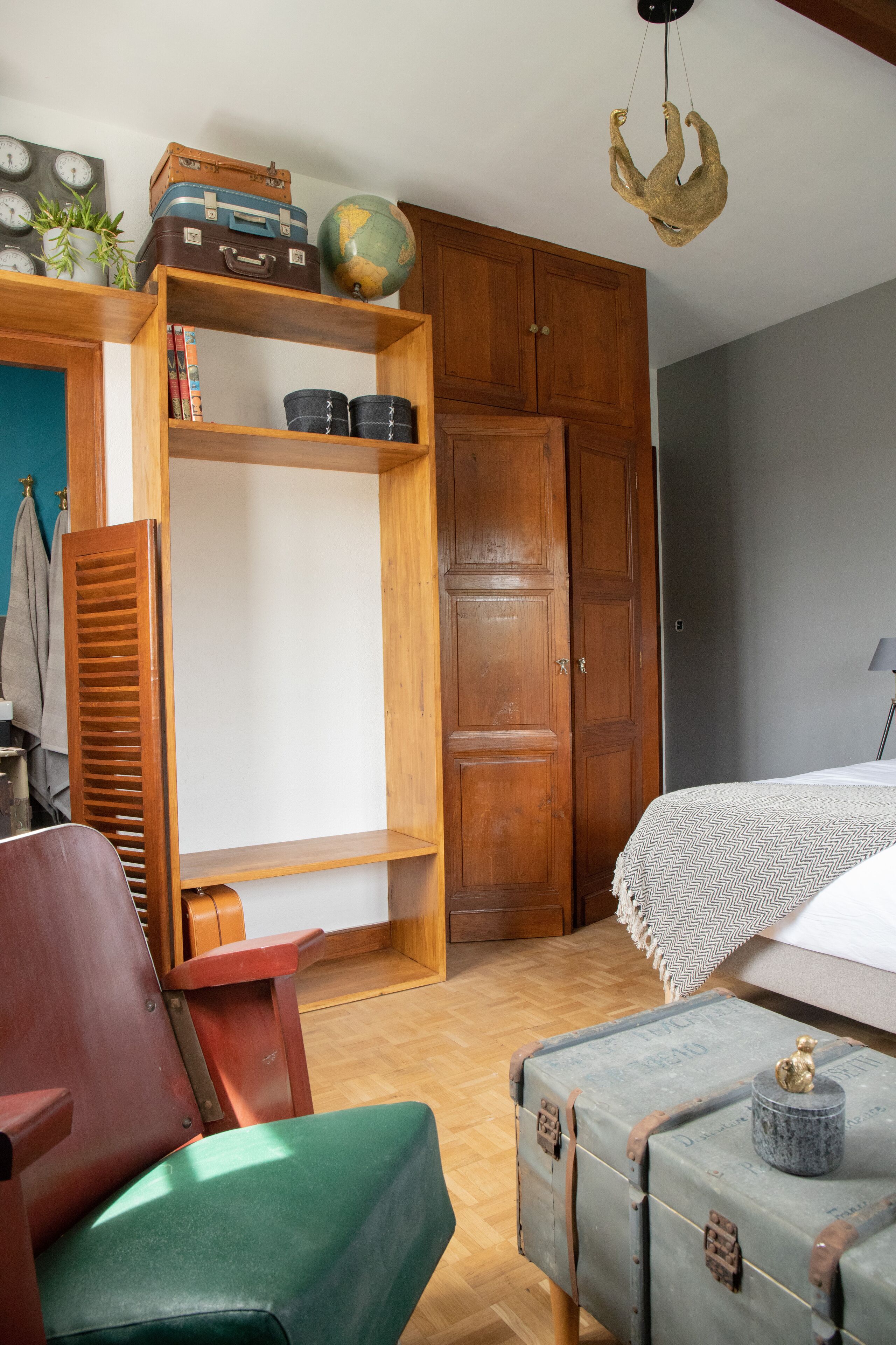 A room with a wooden wardrobe, a green chair, a bed with a patterned bedspread, and a suitcase with a globe on top of a shelf.