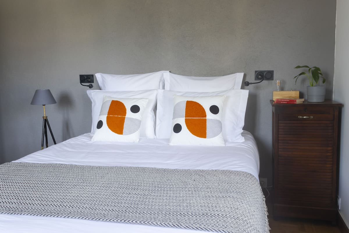 A neatly made bed with white bedding and two pillows featuring orange and black patterns. A gray throw blanket is spread across the bed. A lamp with a gray shade is on a bedside table, and a small plant is on a wooden cabinet beside the bed.