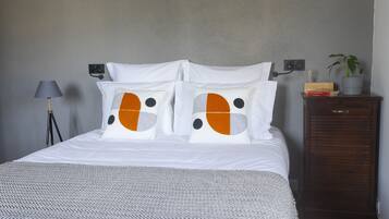 Un lit bien fait, avec une literie blanche et deux oreillers ornés de motifs orange et noirs. Un plaid gris est posé sur le lit. Une lampe avec un abat-jour gris se trouve sur une table de chevet, et une petite plante est placée sur une commode en bois à côté du lit.