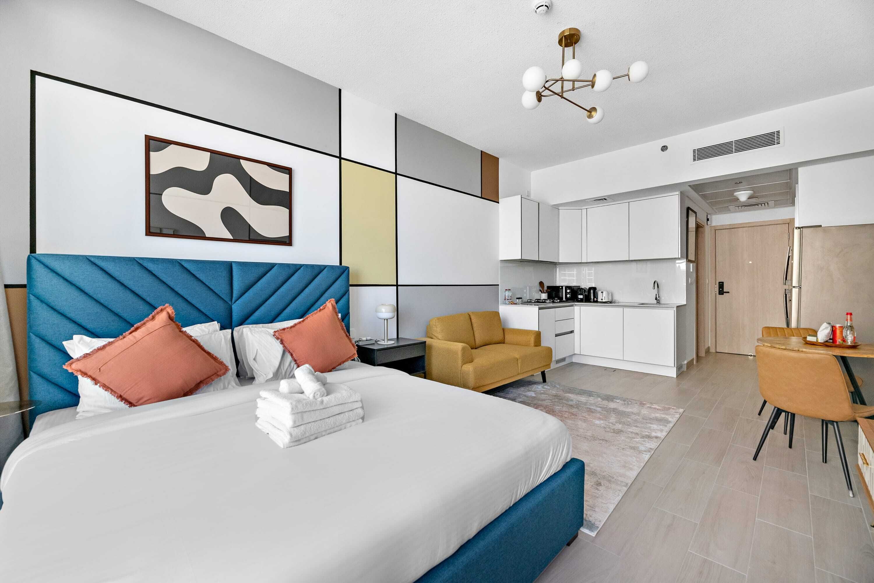 A modern hotel room with a large bed, a desk, a sofa, and a kitchenette.