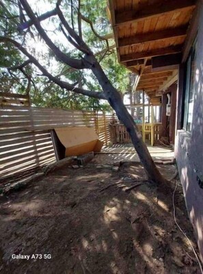 Un espacio exterior techado con una estructura de madera, una valla de metal y un árbol apoyado contra la pared.