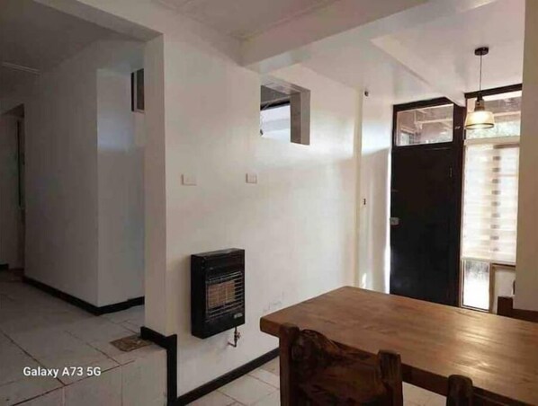 A spacious room with a wooden dining table, a built-in heater, and a door leading to another room.