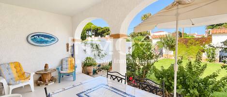 Un patio iluminado por el sol con una mesa, sillas y vistas a un jardín y edificios.