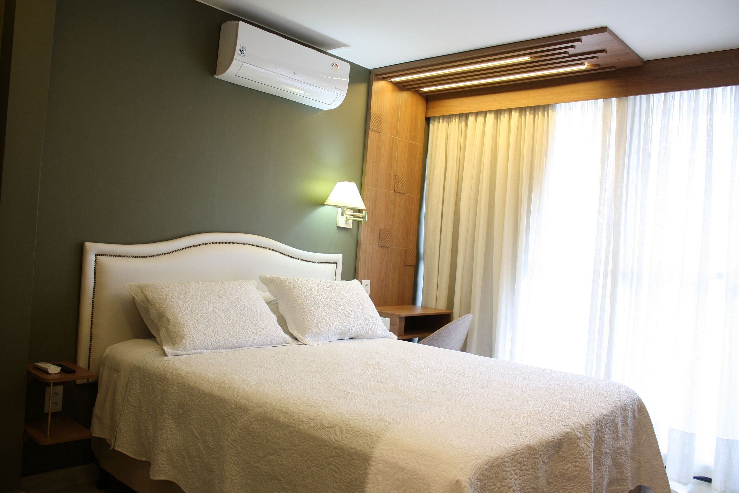 Una cama bien hecha con cubrecama y almohadas blancas, una mesita de noche con lámpara y un equipo de aire acondicionado montado en la pared.