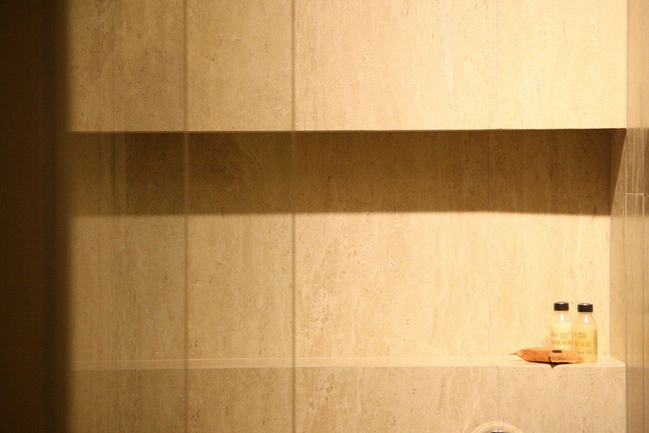 A bathroom with beige tiles and a shelf holding two bottles and a soap bar.