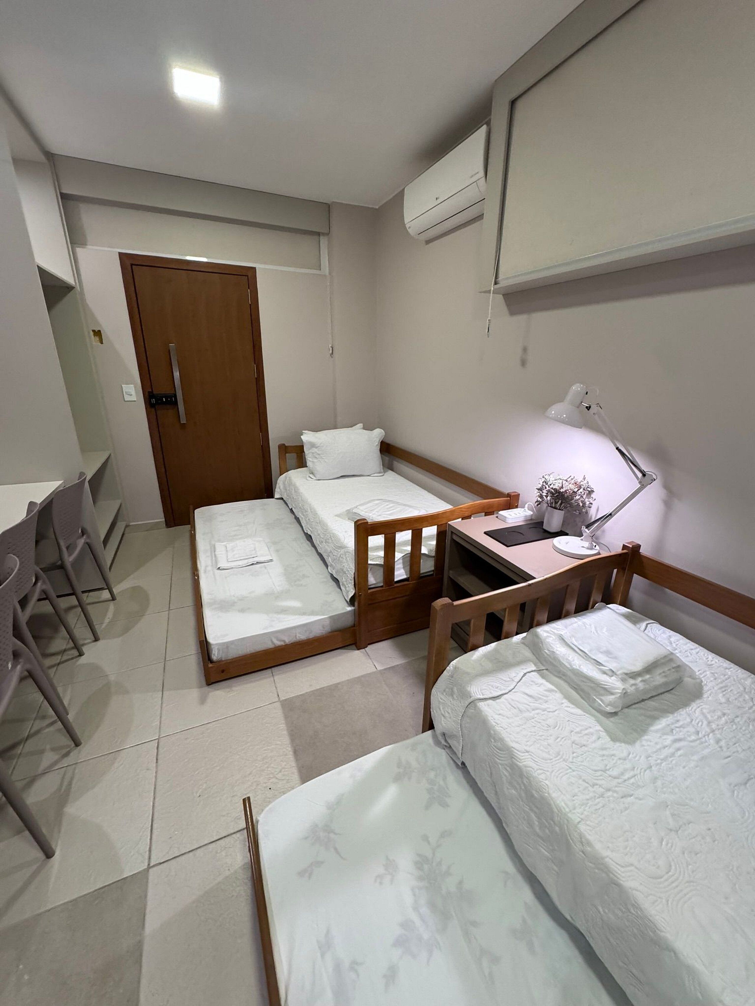 A small, clean room with two beds, a desk, a chair, and an air conditioning unit.
