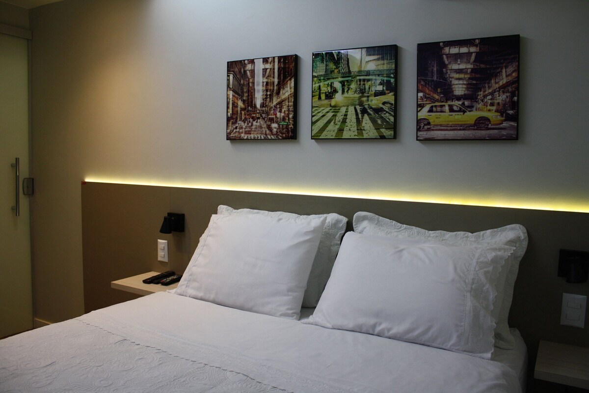 A hotel room with a bed, two white pillows, a nightstand with a phone, and three paintings on the wall.