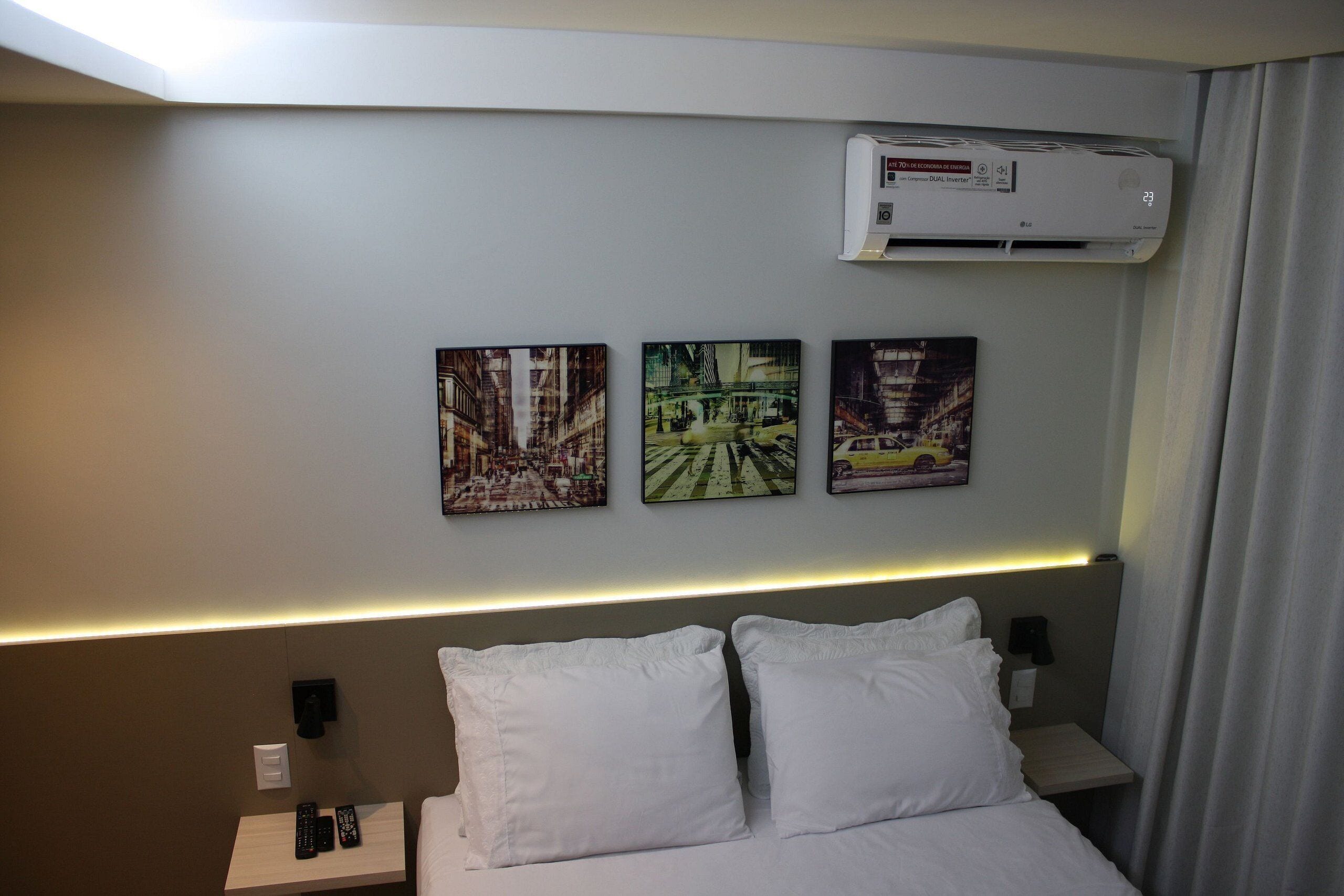 Une chambre d’hôtel avec un lit, des oreillers blancs, une table de chevet avec une télécommande et trois tableaux encadrés accrochés au mur.