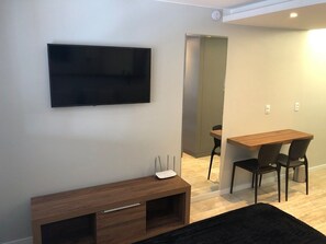 A modern living room with a flat-screen TV mounted on the wall, a wooden TV stand with a white device on top, and a dining area with a wooden table and two chairs. - Flats Asa Norte CLN 110 bloco B (Brasília)