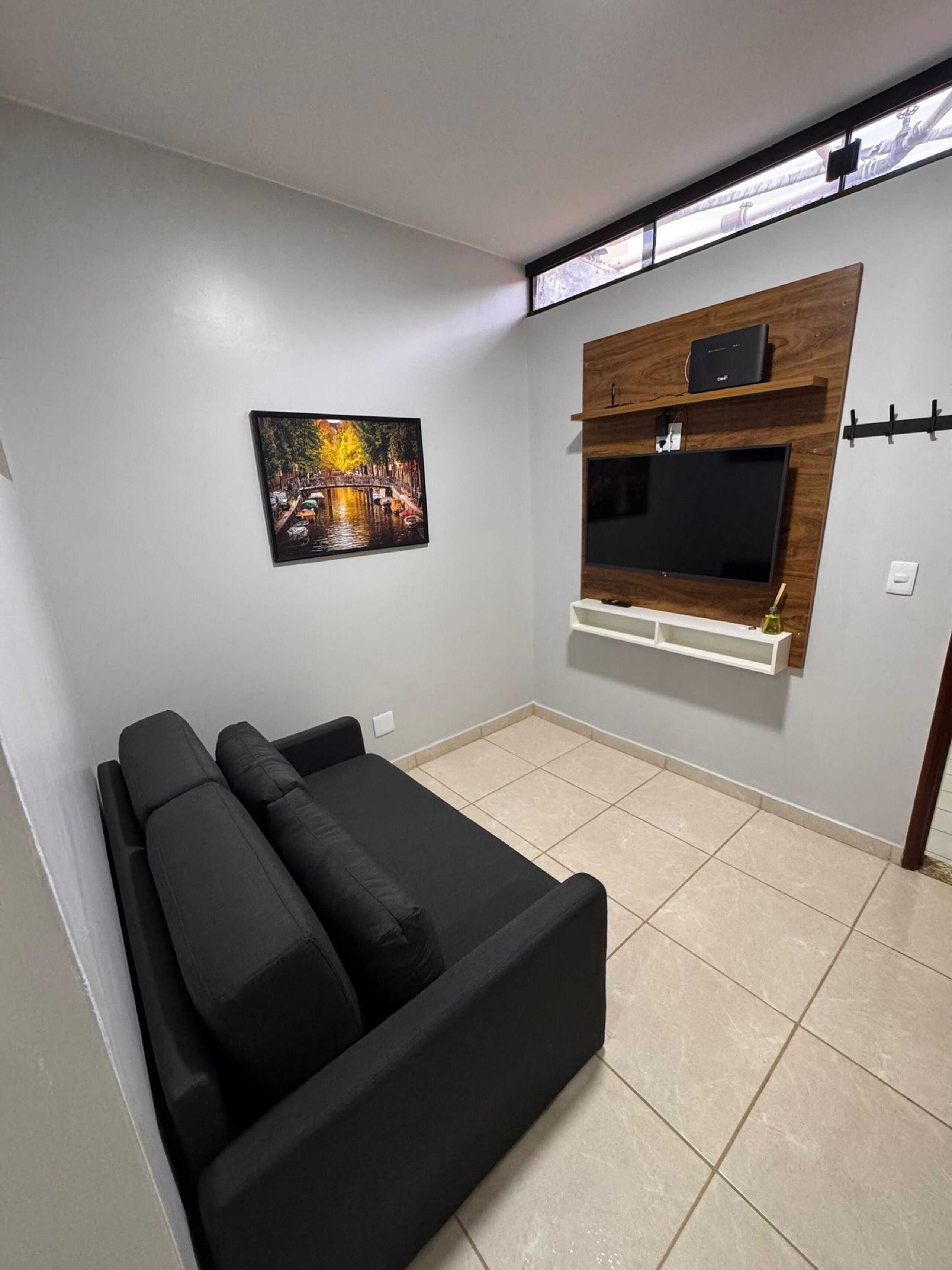 A living room with a black sofa, a mounted TV, a picture of a bridge, and a window with blinds.